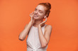 © timtimphoto - Studio photo of good looking young female wearing her red hair in knot, posing over orange background with raised hands to her ears, enjoying music track with closed eyes, wearing white top