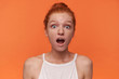 Close-up of surprised young female wearing her red hair in knot over orange background, wearing white top, looking to camera with wide mouth opened and rounding eyes amazedly