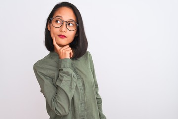 Wall Mural - Young chinese woman wearing green shirt and glasses over isolated white background with hand on chin thinking about question, pensive expression. Smiling with thoughtful face. Doubt concept.