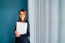 Portrait Of Young Business Woman With Glasses Secretary Holding File Folder Job Application In Front Of Blue Wall By Window Curtains Smiling