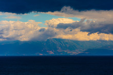 A Mountain Range In Greece Covered In Low Hanging Cloud With The Ocean In The Foreground.