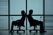 © DragonImages - Silhouettes of two business people sitting back to back in office chairs against big panoramic window and working on laptop