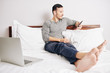 © DragonImages - Barefoot young man resting on bed and reading article on screen of his smartphone