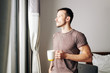© DragonImages - Handsome pensive young man drinking big mug of coffee and looking through window of his bedroom in the morning