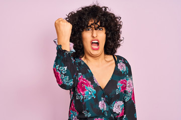 Sticker - Young arab woman with curly hair wearing floral dress over isolated pink background angry and mad raising fist frustrated and furious while shouting with anger. Rage and aggressive concept.