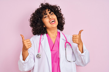 Sticker - Young arab doctor woman with curly hair wearing stethoscope over isolated pink background success sign doing positive gesture with hand, thumbs up smiling and happy. Cheerful expression