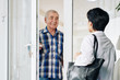 © DragonImages - Smiling senior man in plaid shirt opening glass door to invite caregiver to house