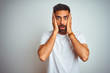 Young indian man wearing t-shirt standing over isolated white background afraid and shocked, surprise and amazed expression with hands on face