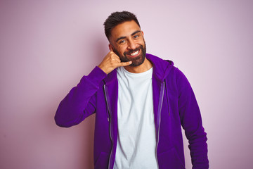 Wall Mural - Young indian man wearing purple sweatshirt standing over isolated pink background smiling doing phone gesture with hand and fingers like talking on the telephone. Communicating concepts.