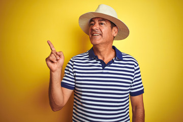 Wall Mural - Handsome middle age man wearing striped polo and hat over isolated yellow background with a big smile on face, pointing with hand and finger to the side looking at the camera.