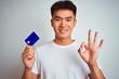 © Krakenimages.com - Young asian chinese man holding credit card standing over isolated white background doing ok sign with fingers, excellent symbol