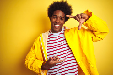 Sticker - African american man with afro hair wearing rain coat standing over isolated yellow background gesturing with hands showing big and large size sign, measure symbol. Smiling looking at the camera