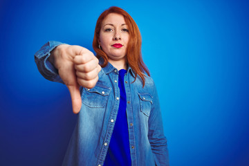 Canvas Print - Young beautiful redhead woman wearing denim shirt standing over blue isolated background looking unhappy and angry showing rejection and negative with thumbs down gesture. Bad expression.