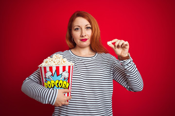 Sticker - Young beautiful redhead woman eating popcorn over red isolated background with a confident expression on smart face thinking serious