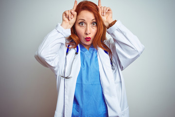 Sticker - Young redhead doctor woman using stethoscope over white isolated background doing funny gesture with finger over head as bull horns