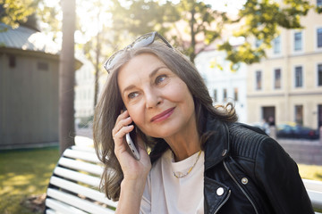 Wall Mural - Close up portrait of fashionable retired female with neat elegant make up spending leisure time outdoors, using cell phone, talking to her son, smiling happily, receiving good positive news