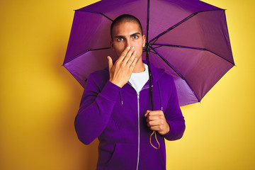 Poster - Young handsome man wearing purple umbrella over yellow isolated background cover mouth with hand shocked with shame for mistake, expression of fear, scared in silence, secret concept