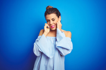 Sticker - Young beautiful woman wearing bun hairstyle over blue isolated background covering ears with fingers with annoyed expression for the noise of loud music. Deaf concept.