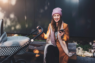 Smiling young woman having day walk in city park. Mother with child in stroller sitting on bench near house. Urban recreation zones for outdoor activities. Female using smartphone for communication.