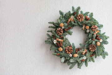 Christmas wreath made of natural fir branches  hanging on a white wall.  Wreath with natural ornaments: bumps, walnuts, cinnamon, cones. New year and winter holidays. Christmas decor. Copy space