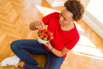 Beautiful young african woman with afro hair eating fresh strawberries sitting on the floor