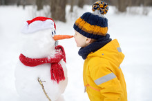 Little Boy Playing With Funny Snowman. Child Reaches For A Snowman's Carrot Nose And Wants To Bite. Active Outdoors Leisure With Children In Winter.