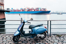 A City Moped Is Parked On The Promenade In Hamburg. In The Background Is Moving A Cargo Ship In The Bay.