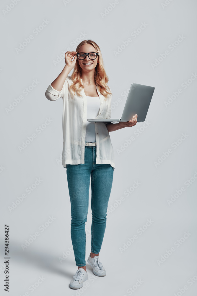 Full length of attractive young woman adjusting eyewear and using laptop while standing against grey background