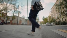 Low Angle Shot Of Young Man In A Blue Pants And White Sneakers With Leather Bag Walking Along The City Crosswalk Alone On Background Of Car Passing And Trees In Autumn Time