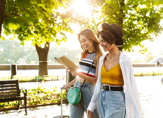 Canvas Print - Photo of two cheerful beautiful women holding map while walking