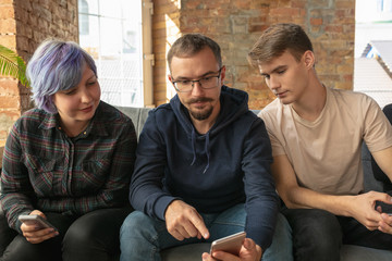 Group of happy young caucasian people sitting on a sofa together. Sharing a news, photos or videos from smartphones, reading articles or playing games and having fun. Social media, modern technologies