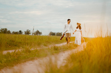 Happy lovers are walking and holding hand on the grassland with tenderness to find a seat to relax. A man with a guitar and a woman with a picnic box.