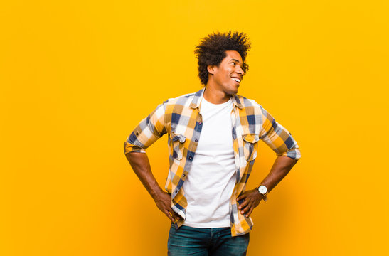 young black man looking happy, cheerful and confident, smiling proudly and looking to side with both hands on hips against orange wall