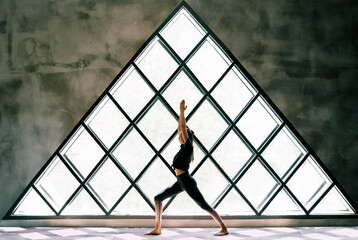 young beautiful woman doing yoga asana warrior i pose on large triangular window background