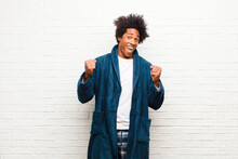 Young Black Man Wearing Pajamas With Gown Feeling Shocked, Excited And Happy, Laughing And Celebrating Success, Saying Wow! Against Brick Wall