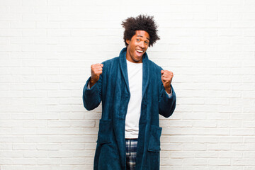 young black man wearing pajamas with gown feeling shocked, excited and happy, laughing and celebrating success, saying wow! against brick wall