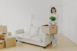 © VK Studio - Full length shot of cheerful brunette woman holds cardboard box with indoor plant, stands in spacious room with sofa and floor lamp, dressed in casual clothes. Female tenant stands in new home