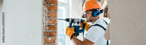 Cuadros en Lienzo panoramic shot of handyman in uniform and yellow gloves using hammer drill
