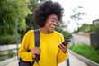 Leinwandbild Motiv smiling young black woman with glasses and bag looking at cellphone outdoors