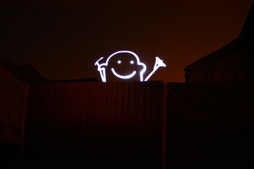 A drawn character man peeks out from behind a fence and smiles, happy smile. Freezelight. Drawing with light in the dark. Shooting at long exposure.