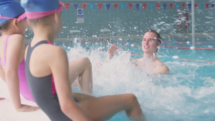 Wall Mural - Children Splashing Male Coach In Swimming Class As They Sit On Edge Of Indoor Pool