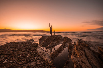 Wall Mural - Hendaye, Aquitaine / France »; October 7, 2019: Photo A photographer on top of some rocks waving at camera in an orange sunset on the beaches of Hendaye