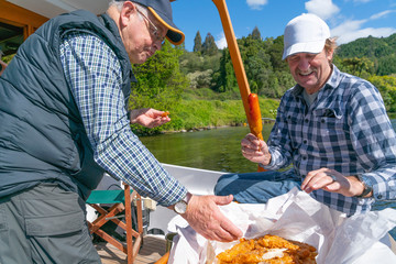 two mature aged men on back of boat on water tucking in to a feed of takeaway fish and chips in paper.