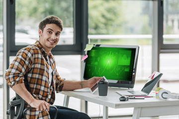 cheerful programmer smiling at camera while sitting near computer monitor with graphs and charts on screen