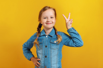 Cheerful little girl shows a victory sign. A child in a denim shirt on a yellow background. Success concept