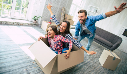 Wall Mural - Family of three is playing in their new apartment after the house moving, young bearded father is riding his wife and daughter in a box, which was just emptied from their personal items.