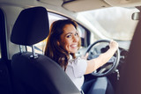 Fototapeta  - Rear view of smiling caucasian brunette dressed smart casual driving her expensive car while looking at camera. Hands are on steering wheel.