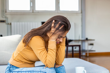 Portrait of a young brunette girl sitting on the couch at home with a headache and back pain. Beautiful woman suffering from chronic daily headaches. Sad woman holding her head because sinus pain