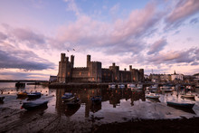 Lit Up Caernarfon Castle At Dusk Reflecting In Bay With Boats In Harbour Long Wispy Clouds Sunset Sky In North Wales