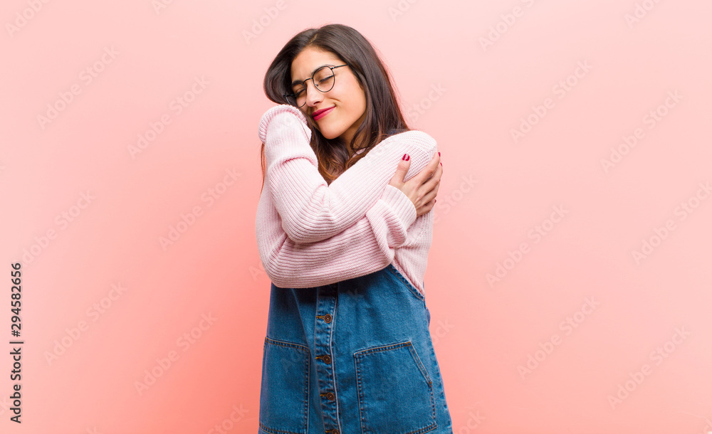 young  pretty woman feeling in love, smiling, cuddling and hugging self, staying single, being selfish and egocentric against pink background. - obrazy, fototapety, plakaty 